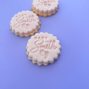 May include: Three round, light pink cookies with scalloped edges. Each cookie has the word "Baby Smith" and two small footprints embossed on the top. The cookies are on a light purple background.