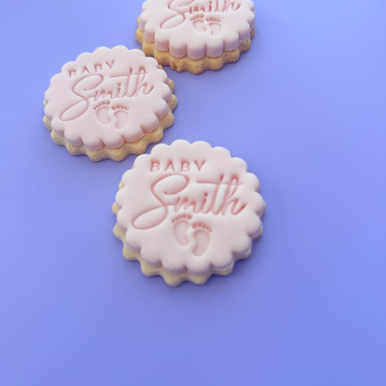 May include: Three round, light pink cookies with scalloped edges. Each cookie has the word "Baby Smith" and two small footprints embossed on the top. The cookies are on a light purple background.