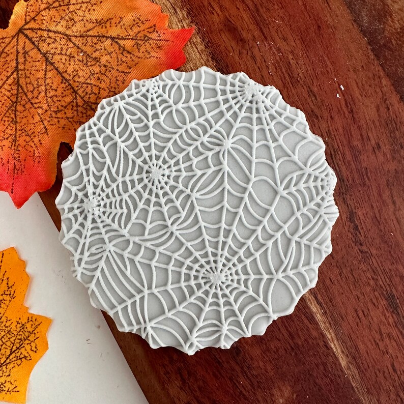 May include: White round cookie decorated with a detailed white spiderweb design.