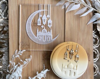 May include: A clear acrylic stamp featuring a crescent moon, mosque, and hanging lanterns design. A round cookie with a similar design is also visible, with gold-colored lanterns and a gradient background. The items are on a wooden surface.