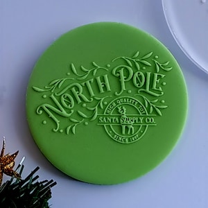 Op de afbeelding: Een rond, heldergroen object met de woorden "North Pole" in een sierlijk lettertype. De tekst "Santa Supply Co. High Quality Toy Since 1897" is ook zichtbaar. Het ontwerp is in reliëf op het oppervlak.