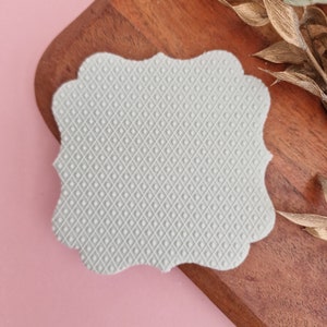 May include: A gray, textured, silicone mold with a decorative, scalloped edge. The mold is shaped like a rectangle with rounded corners.