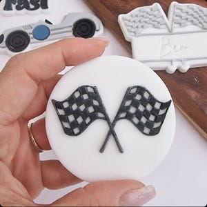 Puede incluir: Una galleta redonda de fondant blanco con un diseño de bandera de carreras a cuadros en blanco y negro. La bandera está cruzada en el centro.