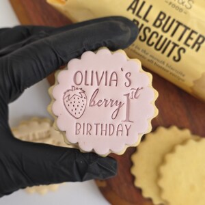 May include: A round, scalloped-edge cookie with pink icing. The icing features the text "OLIVIA'S berry 1st BIRTHDAY" and a strawberry illustration. The cookie is held by a gloved hand, with a blurred background of a biscuit package.