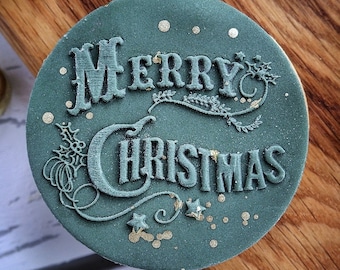 May include: A round, teal-colored Christmas decoration with the words "MERRY CHRISTMAS" embossed in a vintage font. Gold glitter accents the design, including stars and dots. The decoration is set against a wooden background.