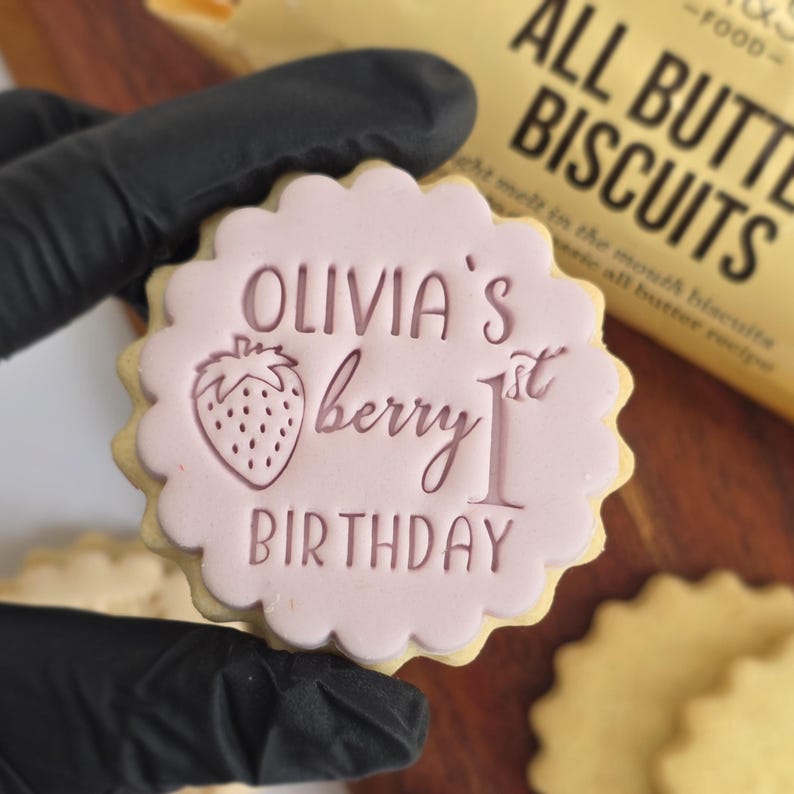 May include: A round, scalloped-edge cookie with pink frosting. The frosting features the text "OLIVIA'S berry 1st BIRTHDAY" and a strawberry illustration. The cookie is held by a gloved hand, with a package of biscuits in the background.
