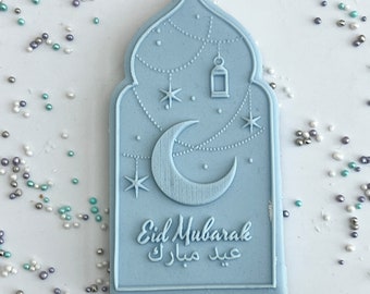 May include: A light blue, arched-shaped decoration with a crescent moon, stars, and a lantern design. The words "Eid Mubarak" are written in English and Arabic. The decoration is surrounded by small, colorful sprinkles.