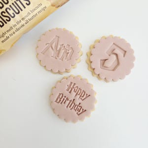 Op de afbeelding: Drie ronde koekjes met roze glazuur en een geschulpte rand. Eén koekje heeft de naam "Ann", een ander het cijfer "5", en de derde zegt "Happy Birthday". Een pakje koekjes is zichtbaar op de achtergrond.
