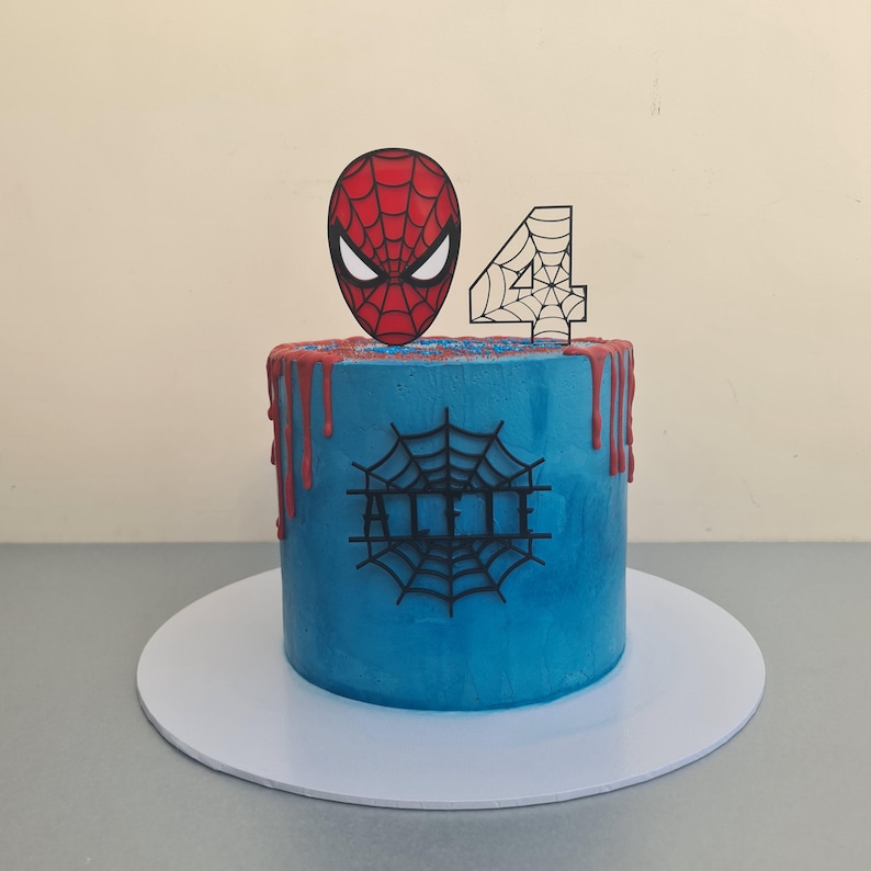 Puede incluir: Un pastel glaseado azul y rojo con una cabeza de Spiderman y un adorno con el n&uacute;mero 4. El pastel est&aacute; decorado con un dise&ntilde;o de telara&ntilde;a negra y el nombre "Allie" escrito en glaseado negro.