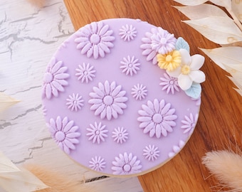 May include: A purple fondant cake decorated with a pattern of small and large flower designs. The cake is topped with a cluster of pastel-colored sugar flowers.