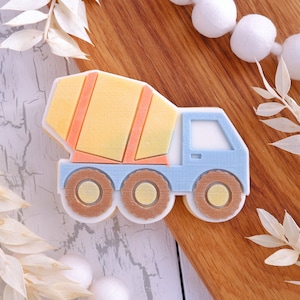 May include: A blue and yellow cookie cutter shaped like a cement mixer truck. The truck has a large, round drum on the back and a smaller cab in the front. The cookie cutter is on a wooden surface.