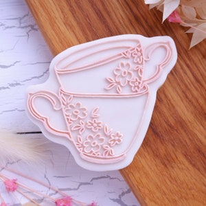May include: A pink and white cookie cutter shaped like a teacup with floral designs. The teacup has a handle and a rim.