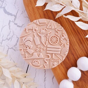 May include: A round, beige silicone stamp featuring a variety of western-themed designs, including a cowboy hat, a cactus, a snake, a wagon wheel, and a wanted poster. The stamp is on a wooden surface.