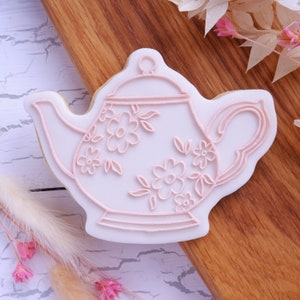 May include: A white cookie decorated with pink icing in the shape of a teapot with floral designs.
