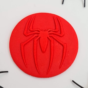 May include: A bright red, circular emblem featuring a raised spider design. The spider has eight legs and a detailed body. The emblem is set against a white background, with black spider leg accents.