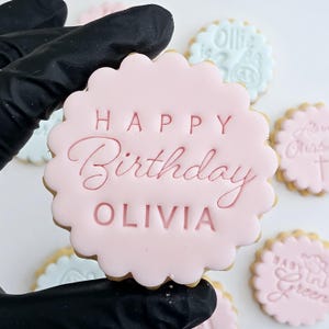 以下が含まれることがあります： 「HAPPY Birthday OLIVIA」の文字が装飾的なフォントで書かれたピンク色のアイシングクッキー。クッキーはスカラップエッジです。背景には文字の書かれた他のクッキーがあります。