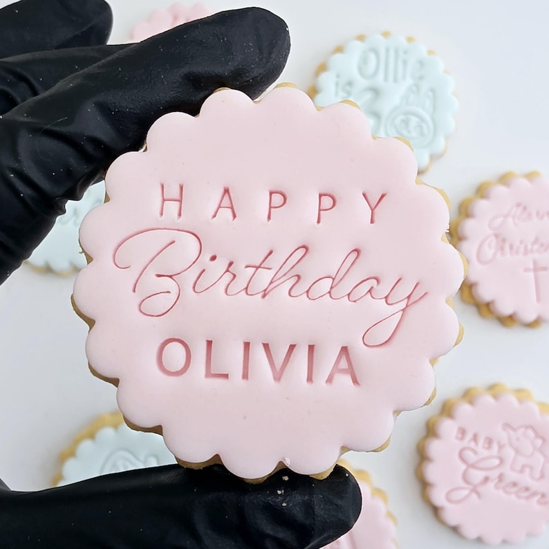 以下が含まれることがあります： 「HAPPY Birthday OLIVIA」の文字が装飾的なフォントで書かれたピンク色のアイシングクッキー。クッキーはスカラップエッジです。背景には文字の書かれた他のクッキーがあります。