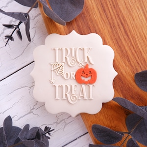 May include: A scalloped-edge biscuit with a smooth off-white surface featuring the embossed text TRICK OR TREAT in a decorative font. The design includes a small white spider web with a hanging spider and a bright orange jack-o'-lantern pumpkin.