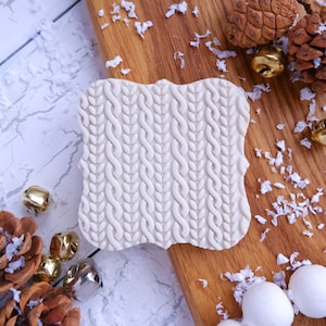 May include: White silicone mould with a raised design of a knitted cable stitch pattern. The mould is on a wooden surface with white paper scraps.