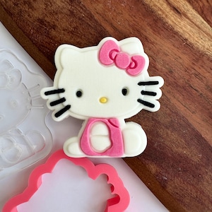 May include: A white fondant Hello Kitty cookie cutter with a pink bow and black whiskers. The cutter is sitting on a wooden surface next to a clear plastic Hello Kitty cookie cutter.