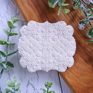 May include: A white, scalloped-edge cookie with an embossed Nordic pattern featuring snowflakes and geometric shapes. The cookie is set on a wooden board, with sprigs of greenery in the background. The design is intricate and detailed.