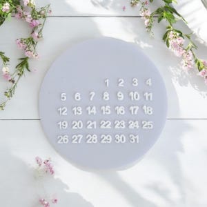 May include: A white calendar with the numbers 1 through 31 printed on a light purple background. The calendar is on a white surface with white flowers.