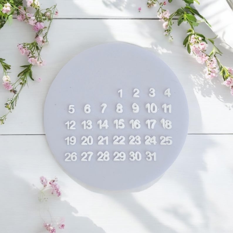 May include: A white calendar with the numbers 1 through 31 printed on a light purple background. The calendar is on a white surface with white flowers.