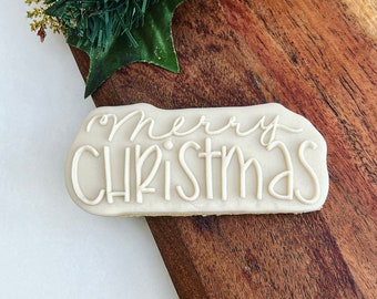 May include: A rectangular cookie with the words "merry CHRISTMAS" in white icing. The cookie sits on a wooden surface, with Christmas greenery and gold accents in the background. A festive holiday treat.