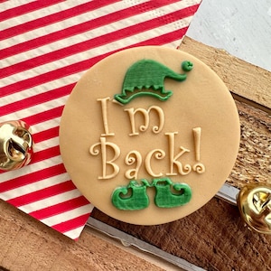 May include: A circular biscuit with the words "I'm Back!" in decorative lettering, accompanied by a green elf hat and shoes. The biscuit is on a wooden surface, with a red and white striped napkin and gold bells.