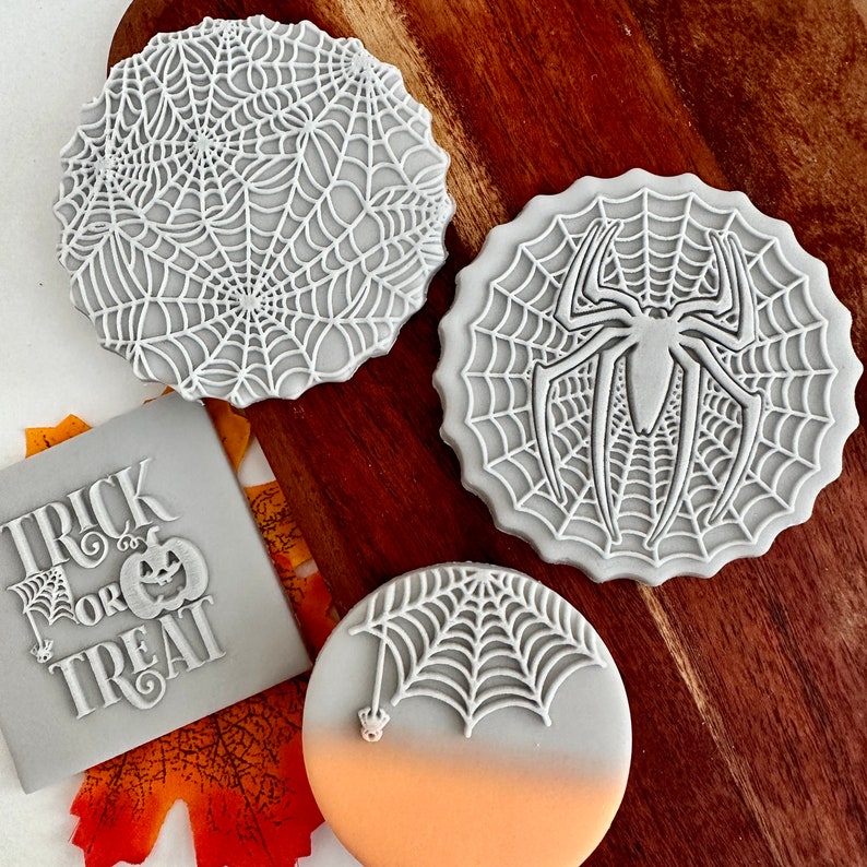 May include: Four gray silicone cookie cutters with Halloween themes. The cutters are shaped like a spider web with a spider, a spider web, a spider web with a spider hanging from it, and a banner that says "Trick or Treat".