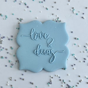 Puede incluir: Una galleta azul claro con las palabras "love & duas" escritas en glaseado blanco.