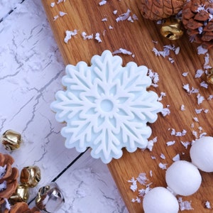 May include: A white snowflake-shaped cookie on a wooden surface with white sprinkles. The cookie is decorated with a light blue glaze.