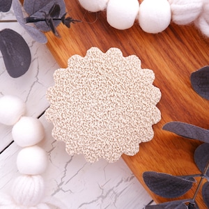 May include: A beige, round, textured coaster with a scalloped edge. The coaster is on a wooden surface with white and grey painted wood in the background.