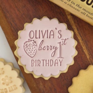 May include: A decorated sugar cookie with a pink frosting design. The cookie features the text "Olivia's berry 1st birthday" and a strawberry illustration. The cookie has a scalloped edge.