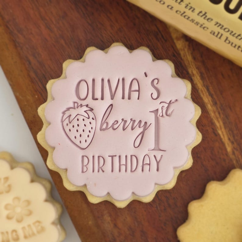 May include: A decorated sugar cookie with a pink frosting design. The cookie features the text "Olivia's berry 1st birthday" and a strawberry illustration. The cookie has a scalloped edge.