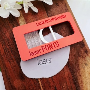 Könnte beinhalten: Eine orangefarbene rechteckige Schablone mit den Worten "LASERCUPBOARD" und "laser FONTS" liegt auf einem grauen, ovalen Objekt mit dem Wort "laser" aufgedruckt. Ein weißes, gebogenes Teil befindet sich in der Schablone.