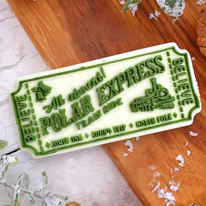 May include: An off-white rectangular Polar Express train ticket with green text and border. The ticket reads "All aboard! POLAR EXPRESS TRAIN RIDE" with a train graphic, and "BELIEVE" on the side. The ticket is on a wooden surface.