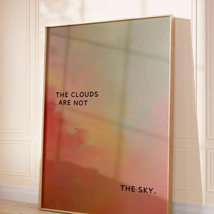 May include: A framed art print with a gradient of colors, including red, orange, and yellow, with the text "THE CLOUDS ARE NOT THE SKY." The frame is a light wood color, and the artwork is leaning against a wall.