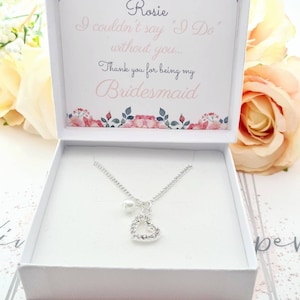 May include: A white gift box with a floral design and the text "Rosie I couldn't say "I Do" without you... Thank you for being my Bridesmaid". The box contains a silver necklace with a heart pendant and a pearl charm.
