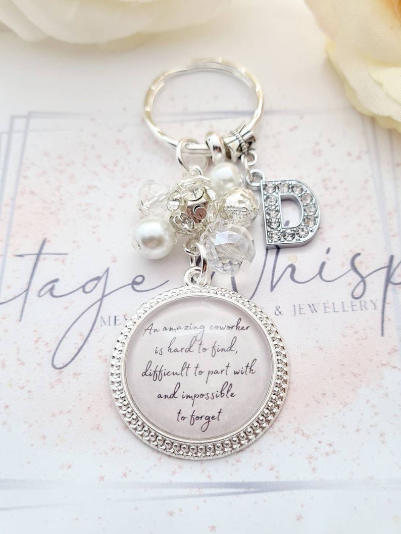 May include: Silver keychain with a charm that reads "An amazing coworker is hard to find, difficult to part with and impossible to forget". The charm is surrounded by white and clear beads and a silver letter D.
