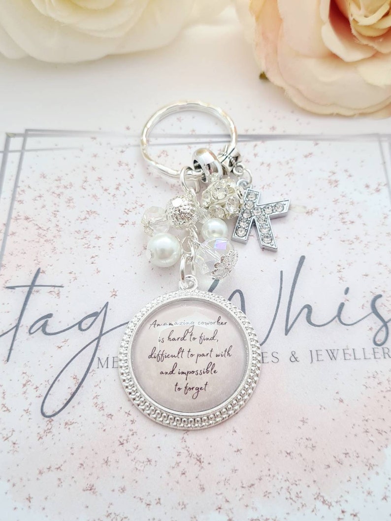 May include: Silver keychain with a charm that reads "An amazing coworker is hard to find, difficult to part with and impossible to forget". The charm is surrounded by small silver beads and a silver letter K.