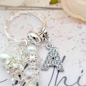 May include: A silver charm keychain with a large letter A charm covered in rhinestones. The keychain also features a variety of other charms, including clear and white beads, and a silver ring.