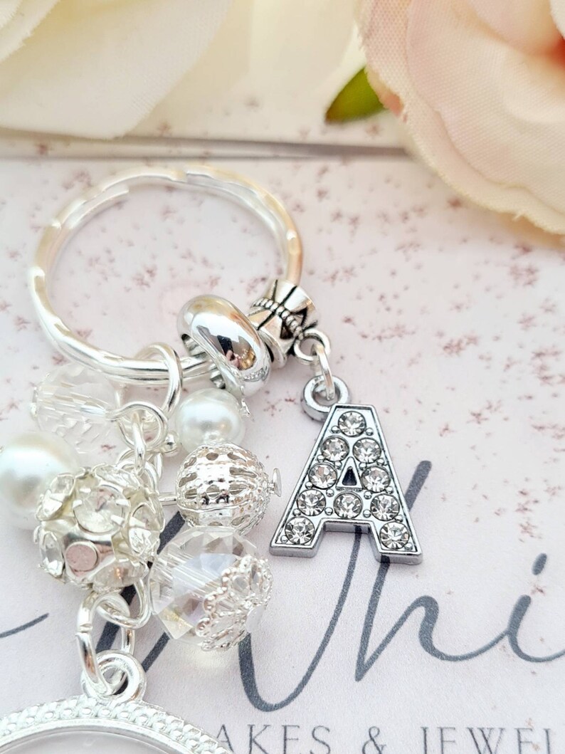 May include: A silver charm keychain with a large letter A charm covered in rhinestones. The keychain also features a variety of other charms, including clear and white beads, and a silver ring.