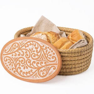 May include: An oval woven basket with a light brown finish containing pieces of bread wrapped in a beige cloth. The basket features a removable terracotta lid with a white embossed floral scroll pattern across the entire surface.