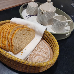 Puede incluir: Una cesta ovalada de mimbre amarillo que contiene varias rebanadas de pan sobre un pa&ntilde;o blanco, junto a una bandeja verde claro con una tetera, un azucarero y una jarra de crema color crema, con un patr&oacute;n de diamantes texturizado.