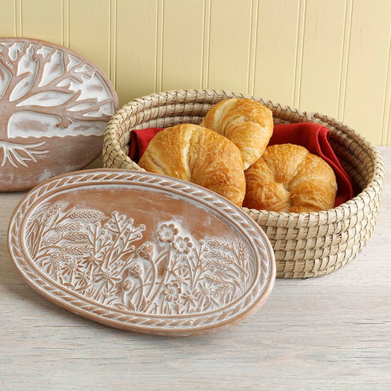 Puede incluir: Una cesta ovalada de mimbre que contiene tres cruasanes dorados sobre un pa&ntilde;o rojo, acompa&ntilde;ada de dos tapas ovaladas de terracota con relieves de flores y &aacute;rboles en blanco, sobre una superficie de madera de color claro.