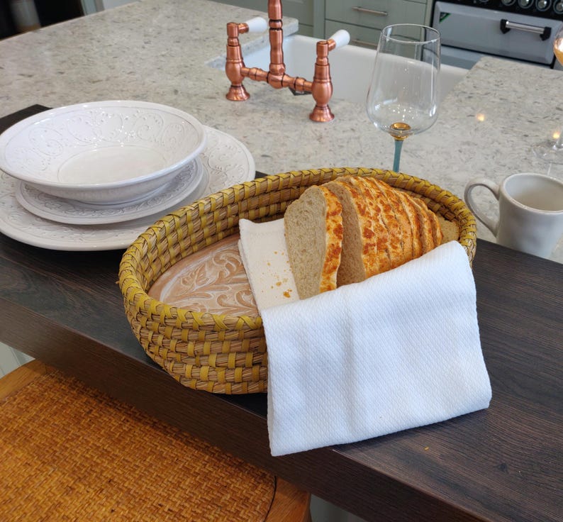 Puede incluir: Una cesta ovalada de mimbre amarillo que contiene varias rebanadas de pan crujiente con queso fundido, parcialmente cubiertas por una servilleta de tela blanca doblada. La cesta tiene una base de madera clara con un dise&ntilde;o de hojas en relieve.