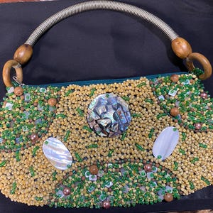 Könnte beinhalten: Eine mit Perlen besetzte Handtasche mit grünem Reißverschluss und Griff. Die Tasche ist mit kleinen, hellgelben Perlen bedeckt und mit grünen Perlen, braunen Perlen und schillernden Muschelteilen akzentuiert. Der Griff ist ein silberfarbener, gewundener Draht mit Holzakzenten.