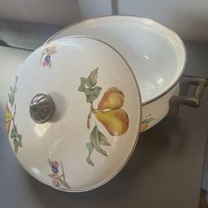 Op de afbeelding: Een witte emaille pan met bijpassend deksel, voorzien van een decoratief patroon van gele peren, groene bladeren en kleine paarse bessen. De pan heeft twee donkere metalen handgrepen aan de zijkanten en een centrale metalen knop op het deksel.