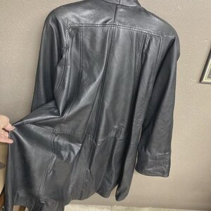 K&ouml;nnte beinhalten: Schwarze Lederjacke mit langer L&auml;nge und klassischem Design. Die Jacke hat einen Kragen, lange &Auml;rmel und einen Knopfverschluss. Das Leder scheint weich und geschmeidig zu sein, mit sichtbaren Nahtdetails.